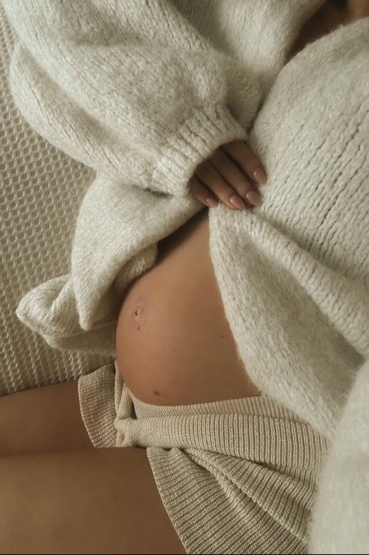 woman wearing a cozy cream maternity sweater, revealing her baby bump while sitting on a textured surface