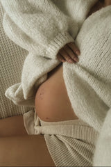 woman wearing a cozy cream maternity sweater, revealing her baby bump while sitting on a textured surface