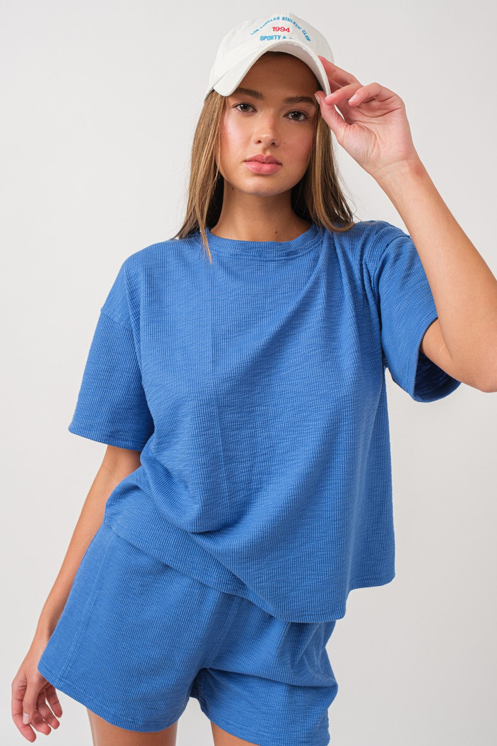woman wearing a light blue short sleeve textured top and matching shorts, holding a white cap with text