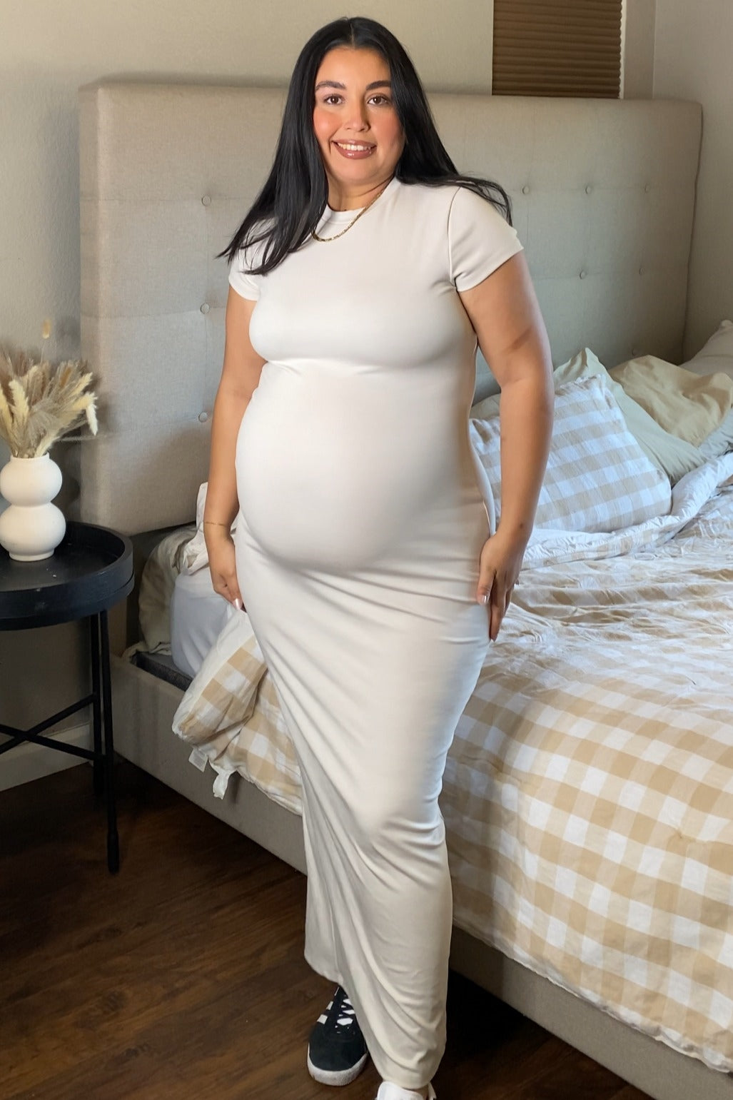 woman wearing a light beige short sleeve maternity maxi dress and showing her baby bump, standing beside a bed