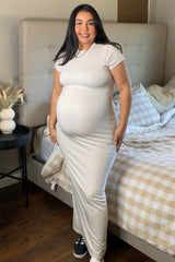 woman wearing a light beige short sleeve maternity maxi dress and showing her baby bump, standing beside a bed