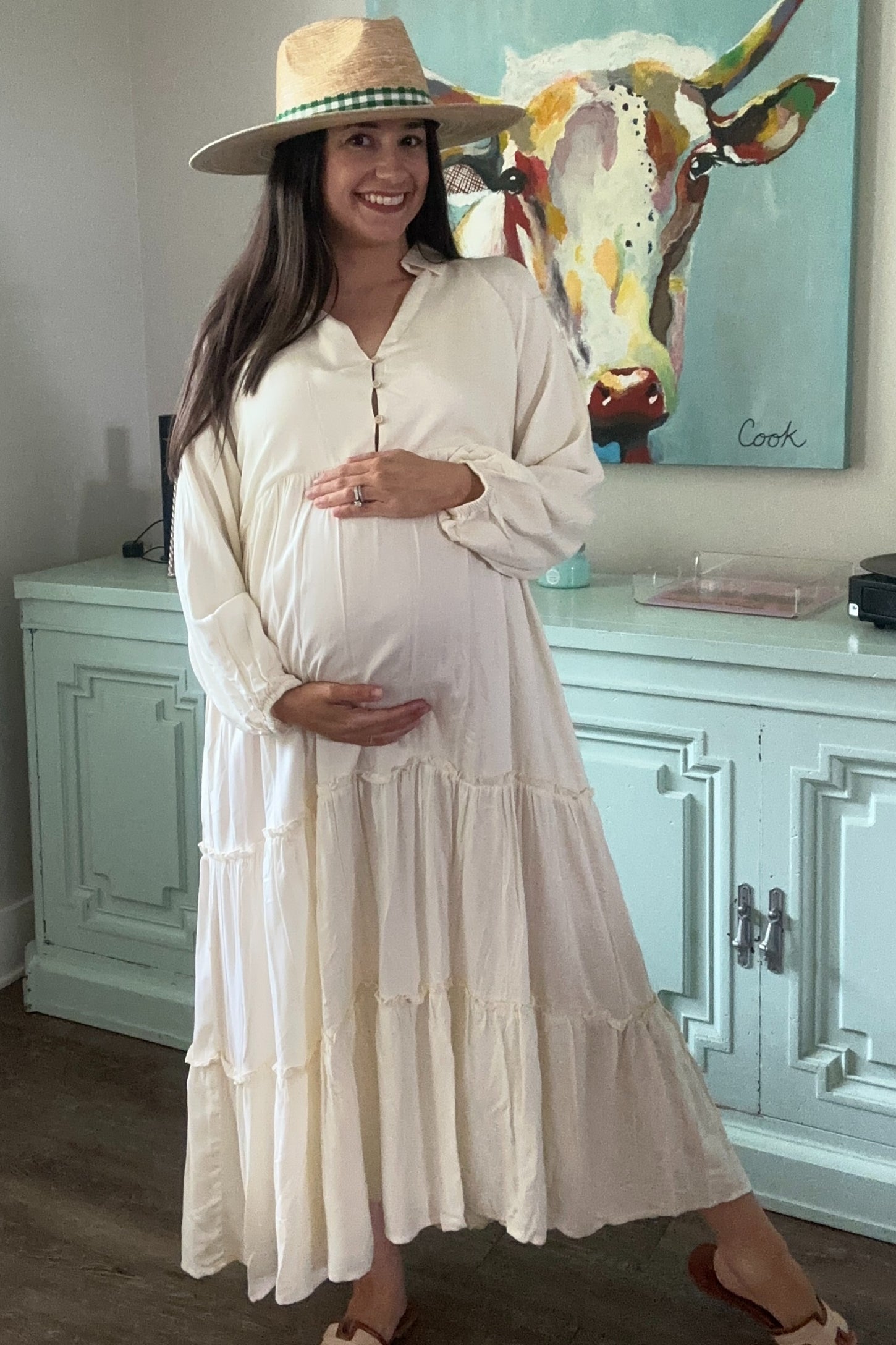 woman wearing a cream long sleeve maternity dress with tiered ruffles and a baby bump, accessorized with a straw hat