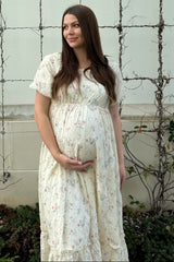 woman wearing a cream floral maternity dress, cradling her baby bump outdoors