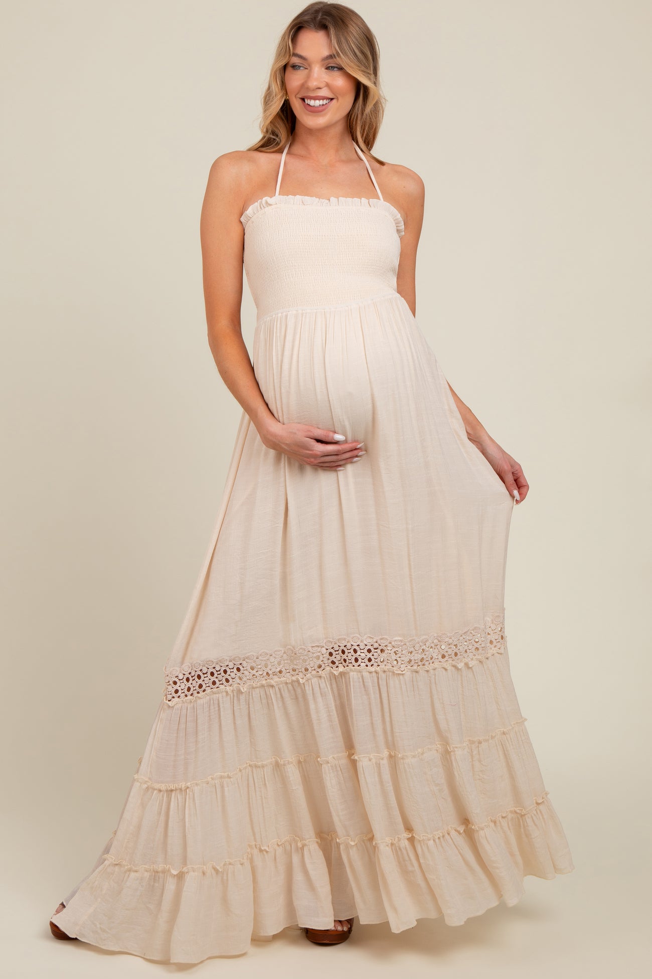 woman wearing a light beige maternity maxi dress with a smocked bodice and ruffled hem, holding her baby bump