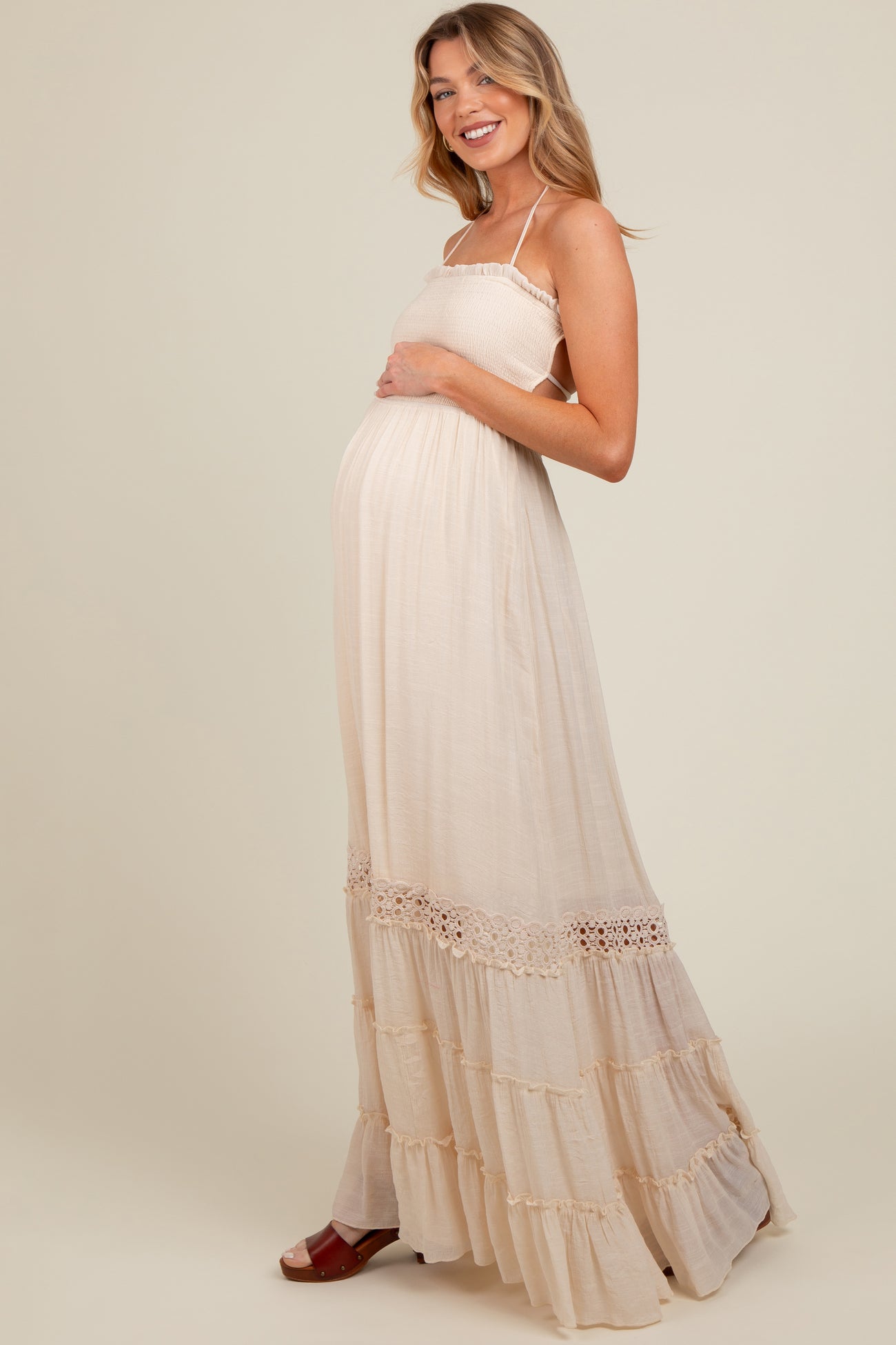 woman wearing a light beige maternity maxi dress with a smocked bodice and ruffled hem, holding her baby bump
