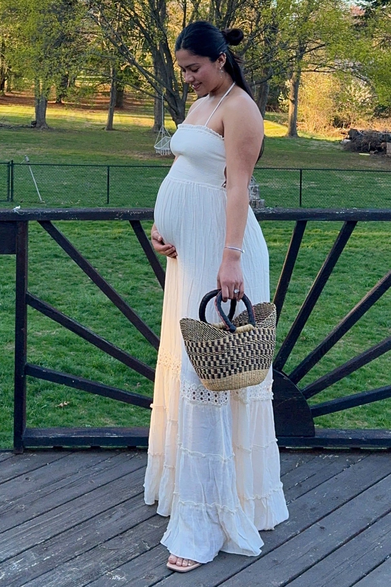 woman wearing a white strapless maxidress, holding her baby bump, and carrying a woven bag
