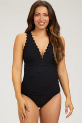 woman wearing a black maternity one-piece swimsuit with scalloped edges and a plunging neckline, displaying her baby bump