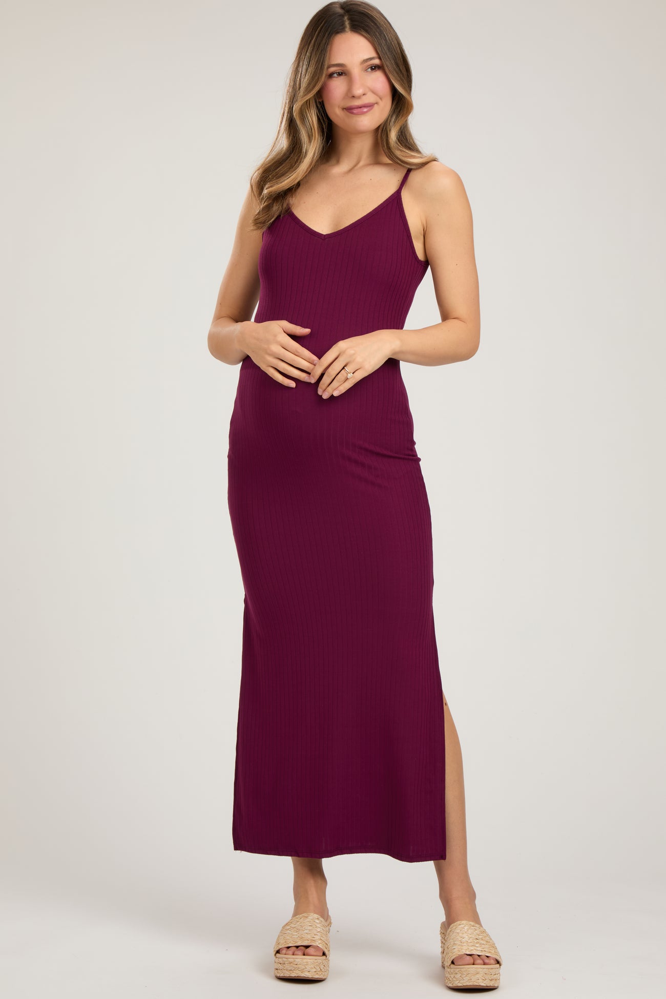 woman wearing a plum maternity maxi dress with a fitted silhouette and side slit, gently holding her baby bump