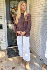 woman wearing a brown polka dot maternity top and white wide-leg jeans, gently cradling her baby bump outdoors