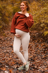 woman wearing a rust oversized sweater and beige leggings, showcasing her baby bump in a fall outdoor setting