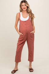woman wearing a rust-colored maternity jumpsuit with buttons and pockets, styled with a white tank top underneath