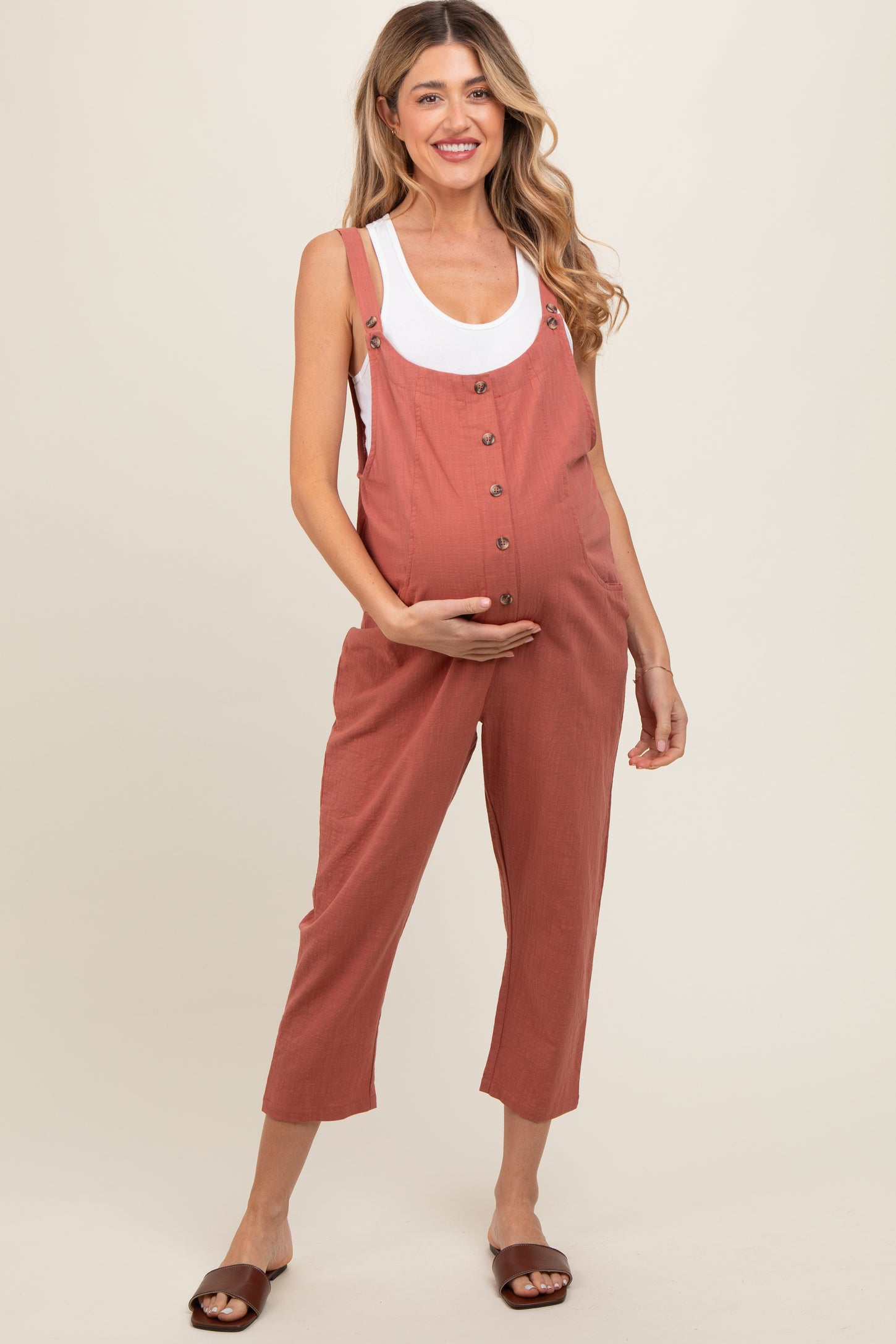 woman wearing a rust-colored maternity jumpsuit with buttons, holding her baby bump and smiling