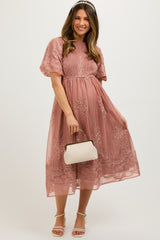 woman wearing a dusty pink maternity dress with floral embroidery and short sleeves, holding a small handbag and showing her baby bump