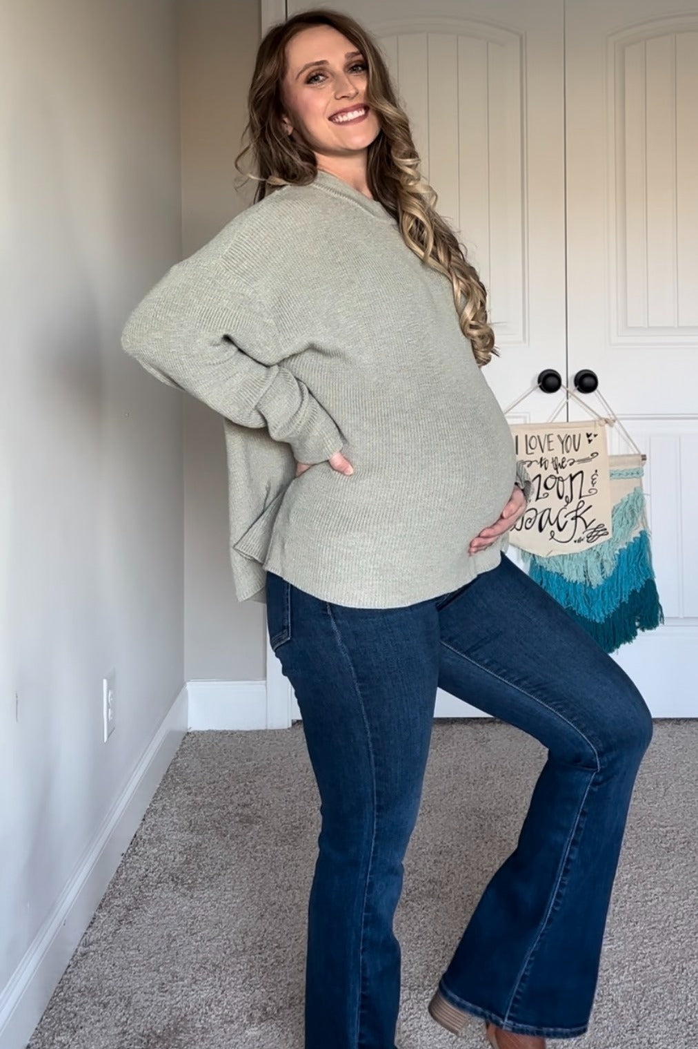 woman wearing a soft green oversized maternity sweater and dark blue flared jeans, showing her baby bump