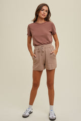 woman wearing a brown short sleeve top and beige drawstring shorts, standing with hands in pockets