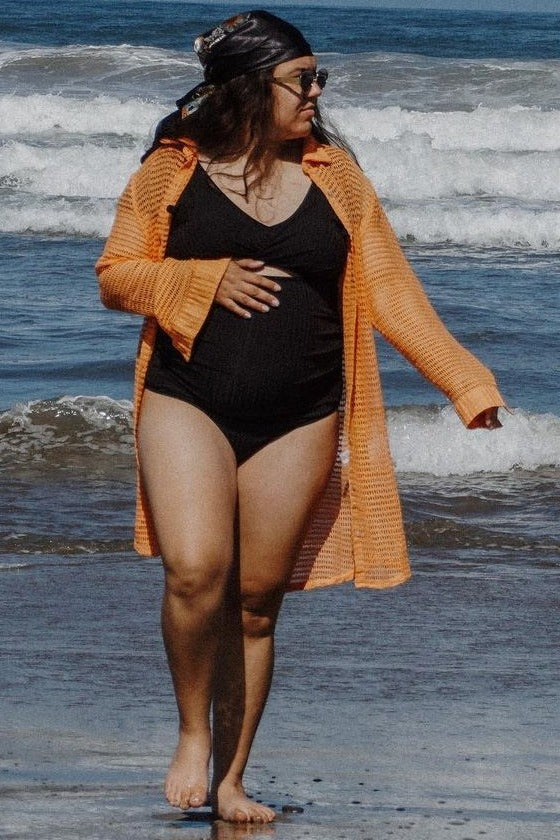 woman wearing a black maternity swimsuit and an orange cover-up, walking on the beach with a visible baby bump