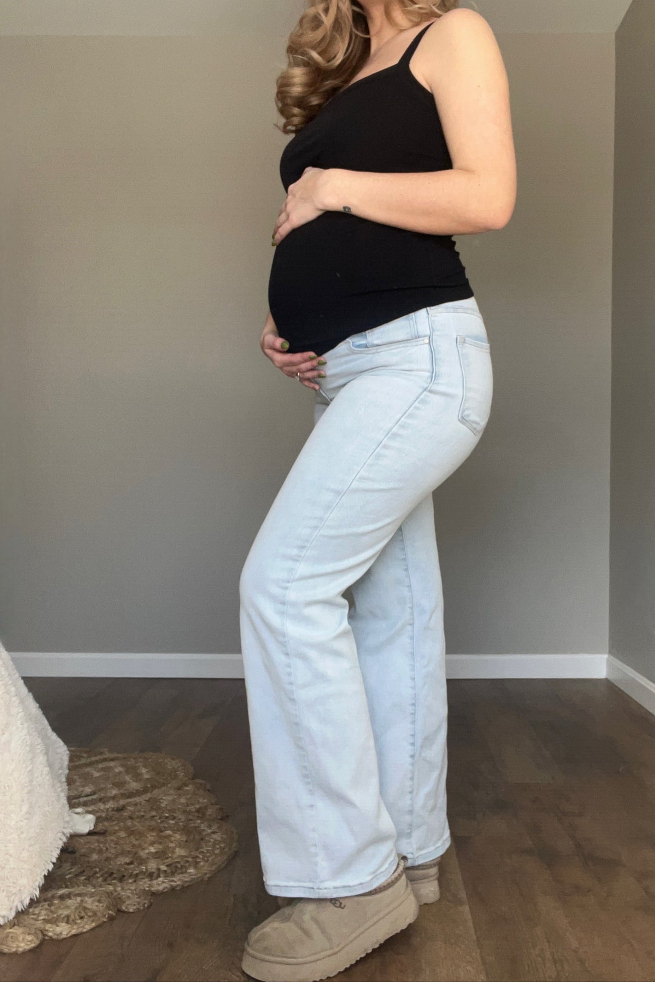 woman wearing a black tank top and light wash maternity jeans, showing her baby bump while posing sideways