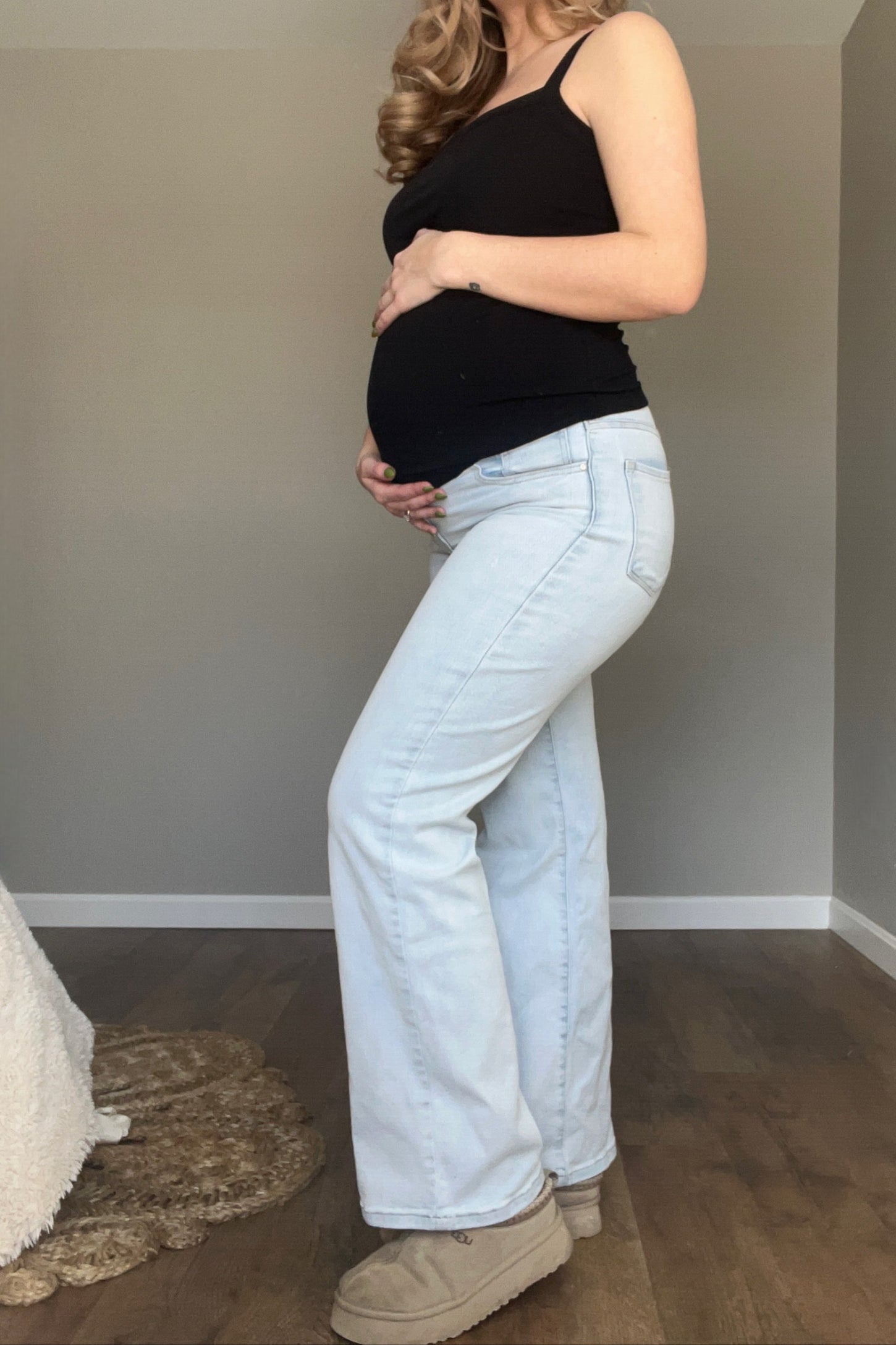 woman wearing a black tank top and light wash maternity jeans, showing her baby bump while posing sideways