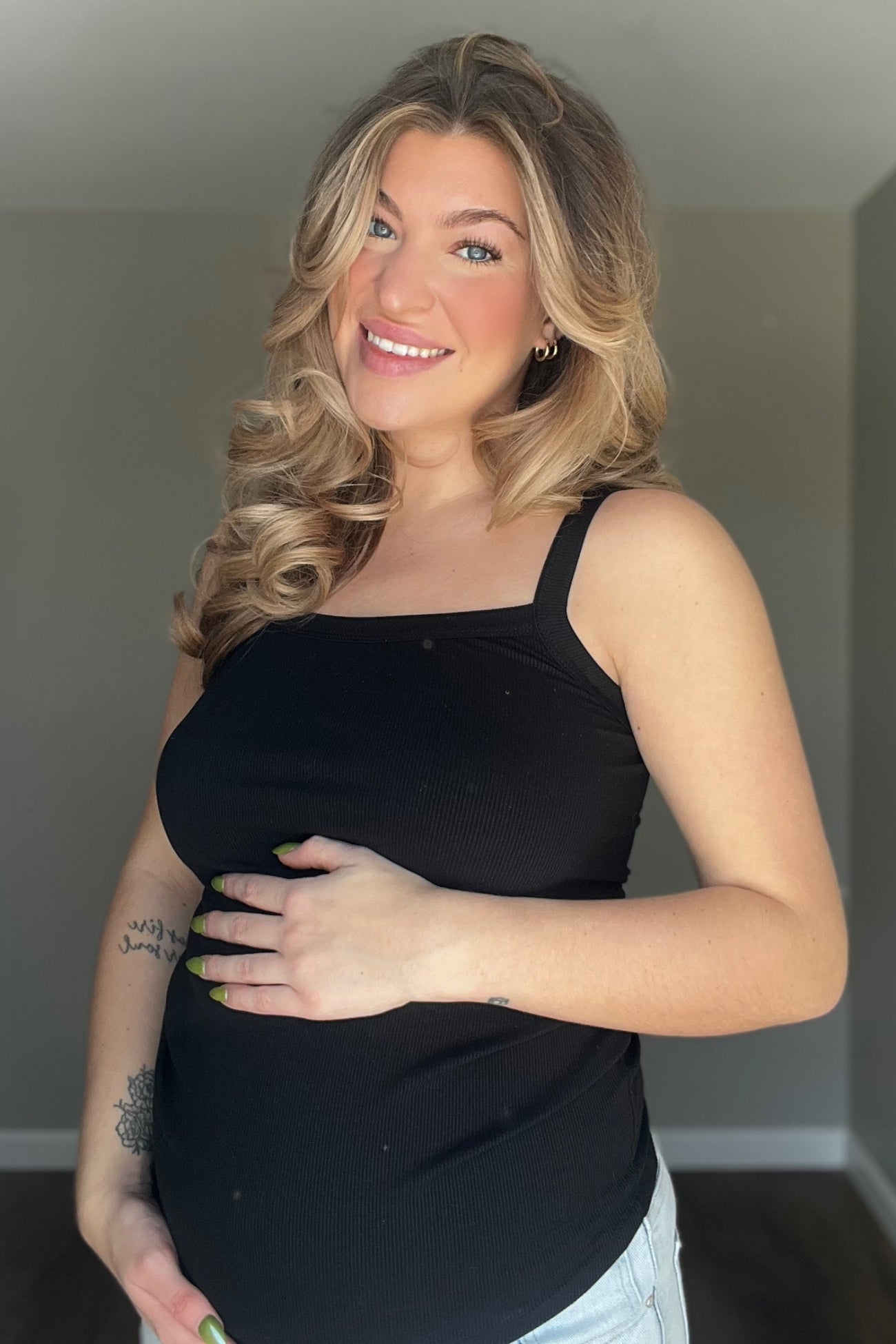 woman wearing a black maternity tank top and holding her baby bump, smiling at the camera