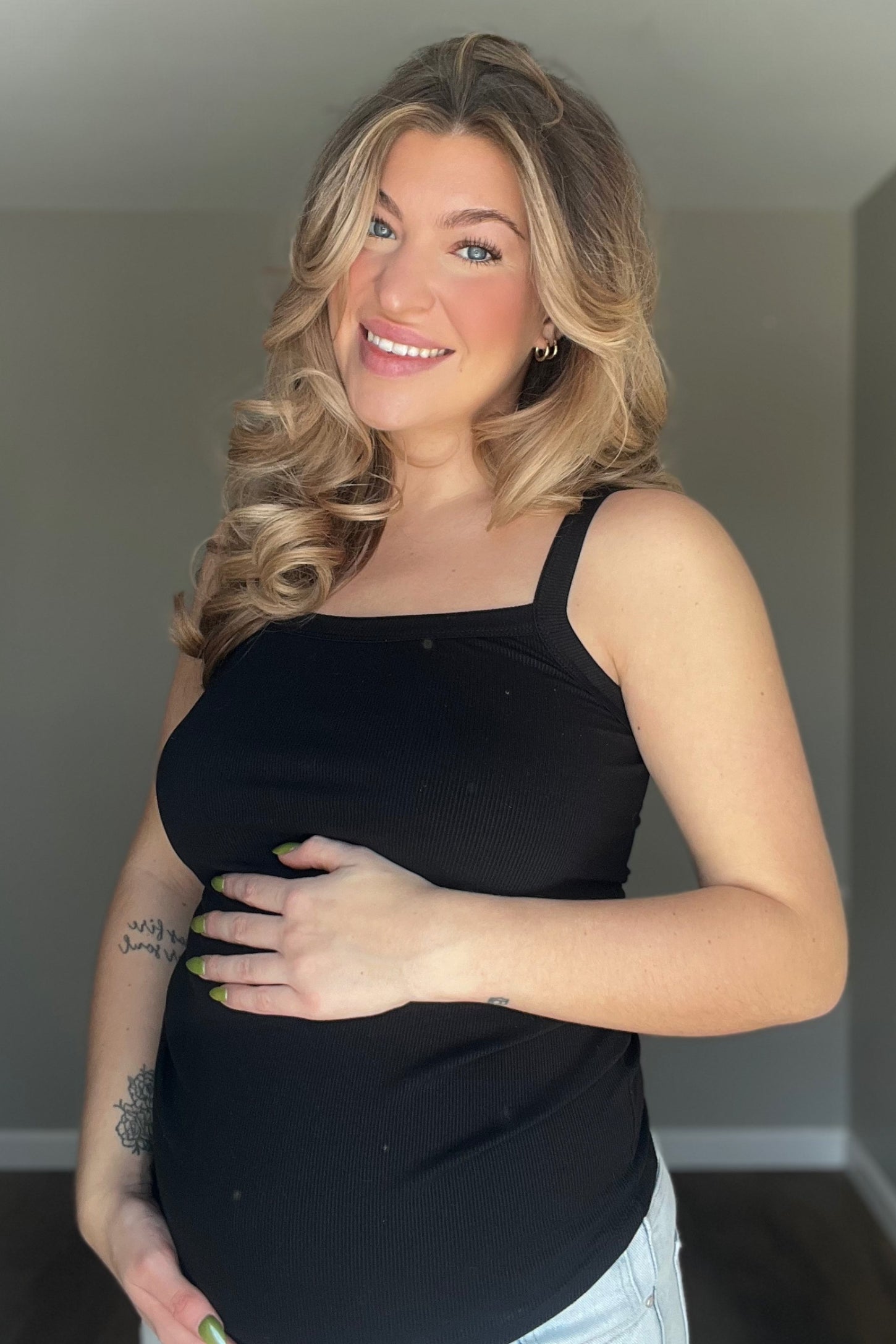 woman wearing a black maternity tank top and holding her baby bump, smiling at the camera