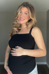 woman wearing a black maternity tank top and holding her baby bump, smiling at the camera