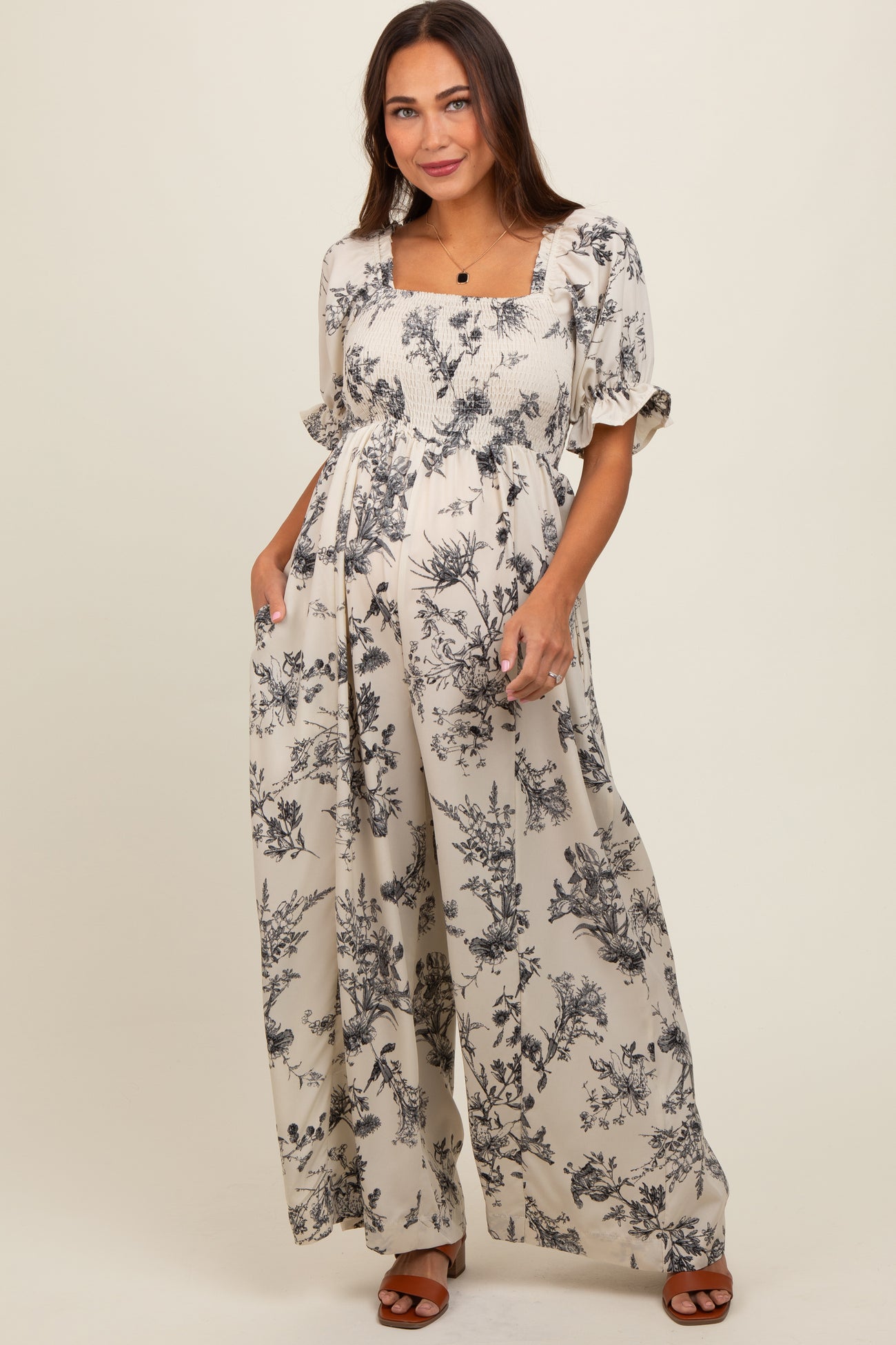 woman wearing a beige floral print maternity jumpsuit with smocked bodice and flowy wide legs, showcasing her baby bump