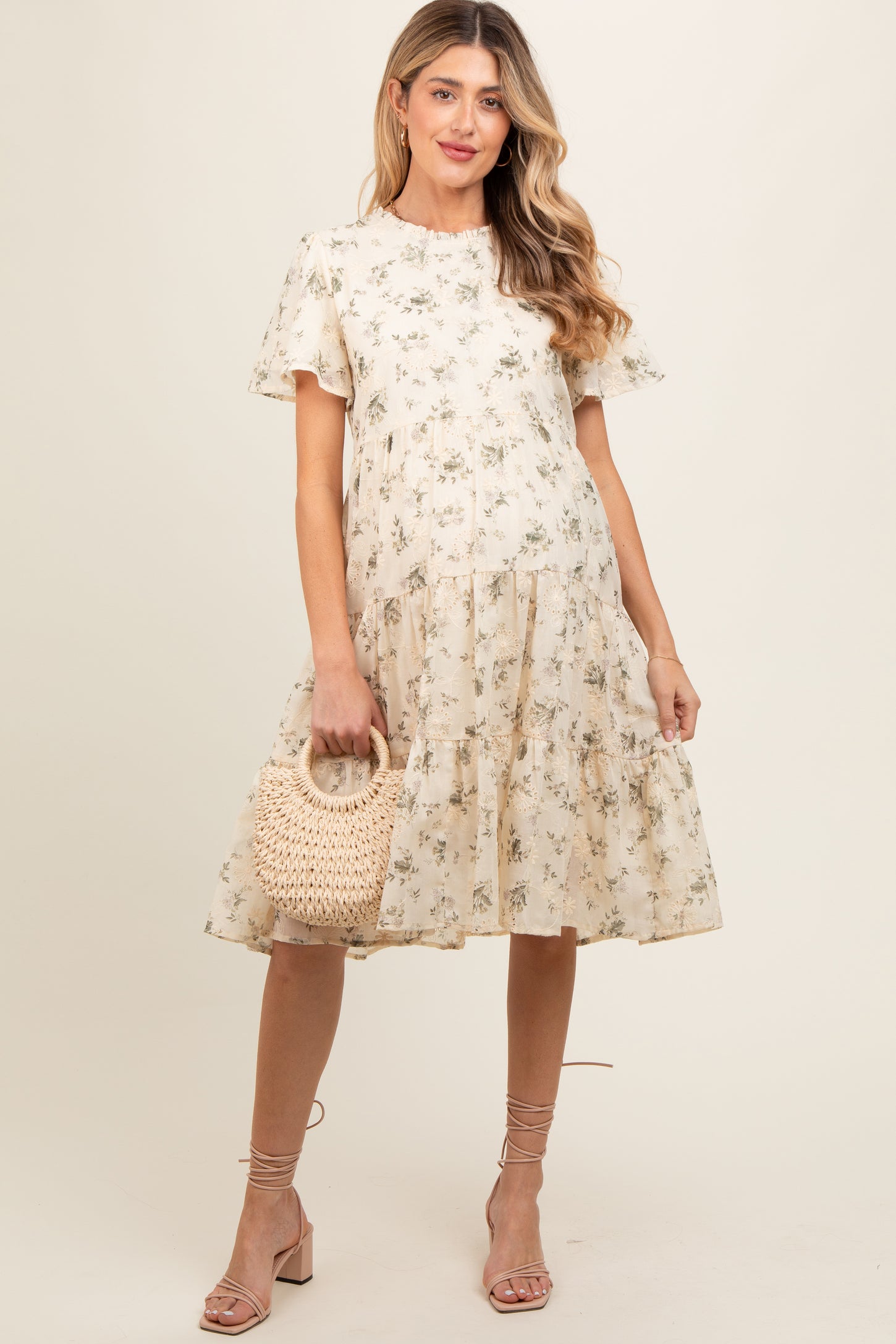 woman wearing a beige floral maternity dress with short sleeves and a tiered skirt, holding a woven bag