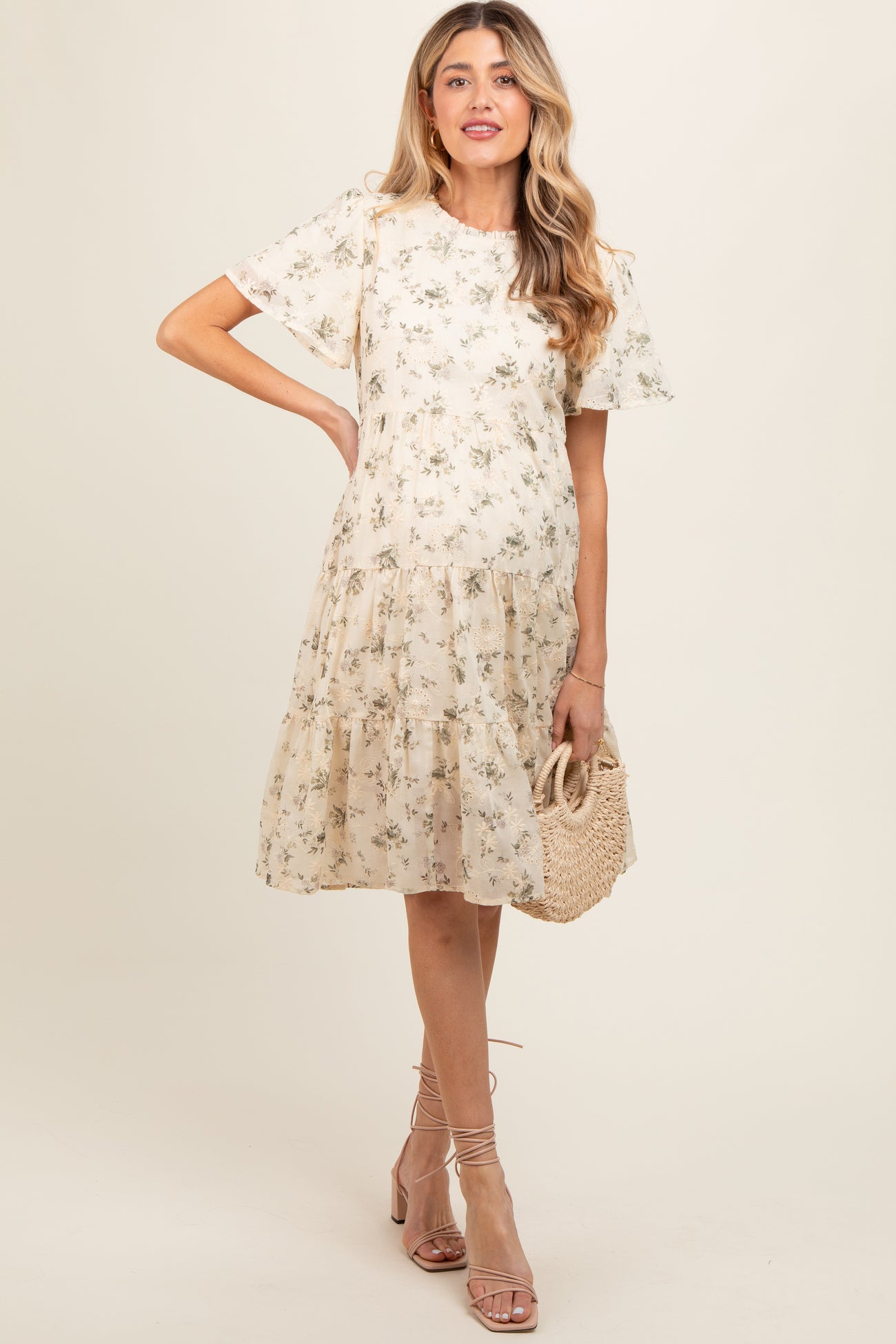 woman wearing a floral print beige maternity dress with short sleeves and a tiered skirt, holding a woven bag