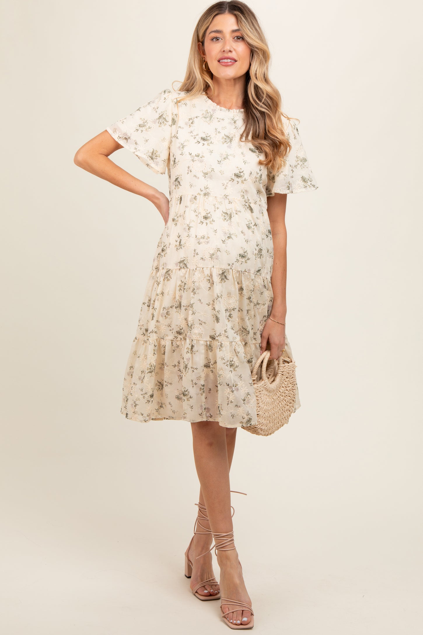 woman wearing a floral print beige maternity dress with short sleeves and a tiered skirt, holding a woven bag