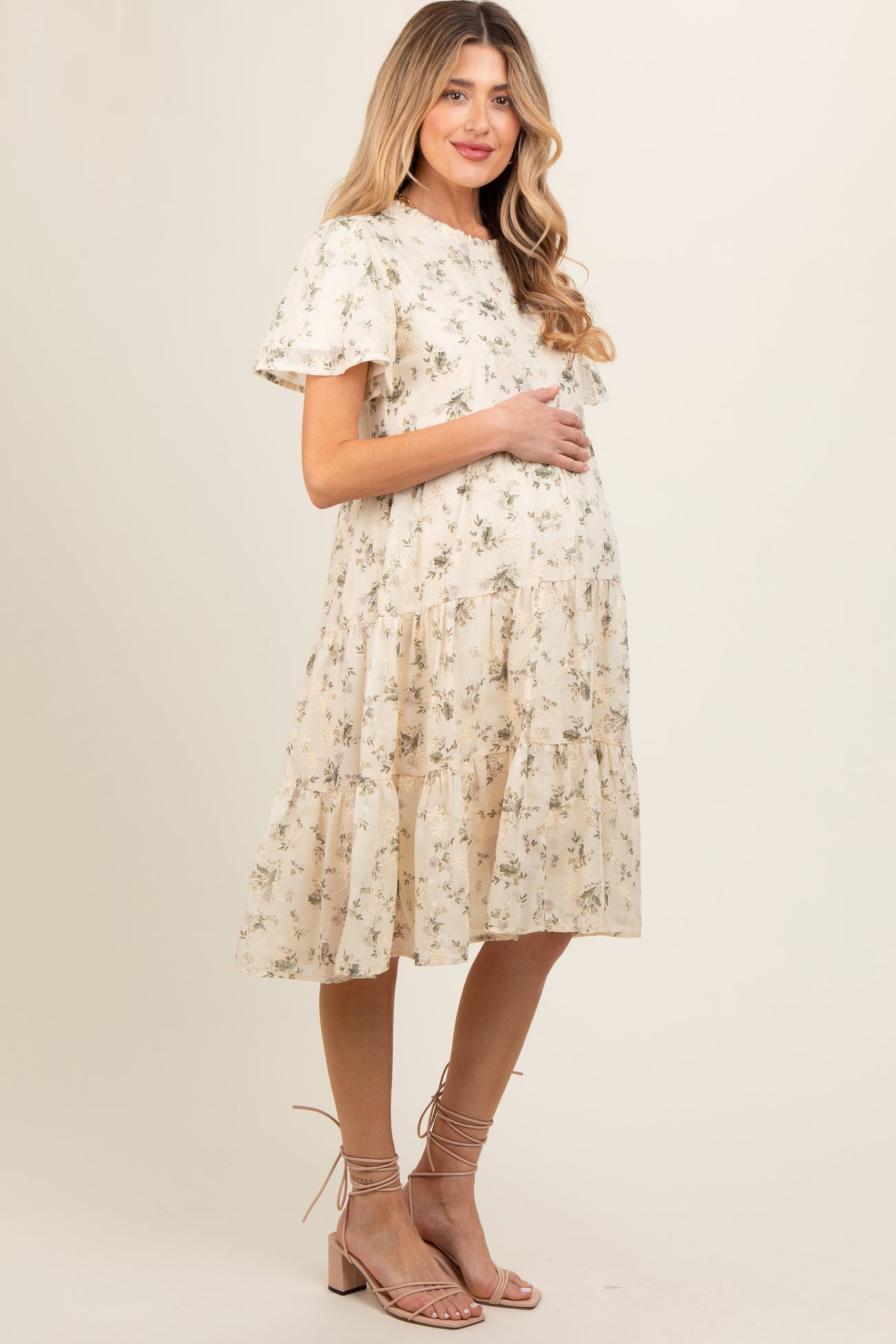woman wearing a cream floral print maternity dress with short sleeves and a tiered skirt, holding her baby bump