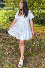 woman wearing a white tiered short sleeve dress and beige sandals, standing in a grassy area with trees in the background