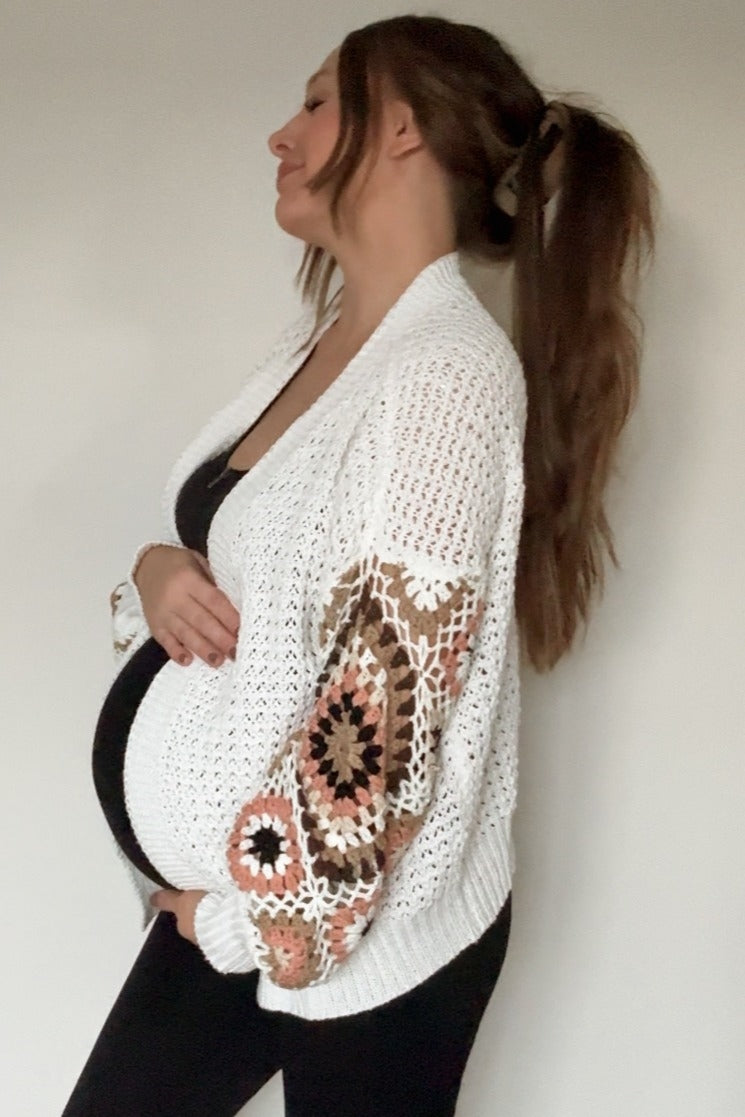 woman wearing a white open-knit maternity cardigan with colorful patterned sleeves and holding her baby bump