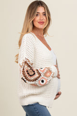 woman wearing a cream crochet maternity cardigan with floral embroidered sleeves and holding her baby bump
