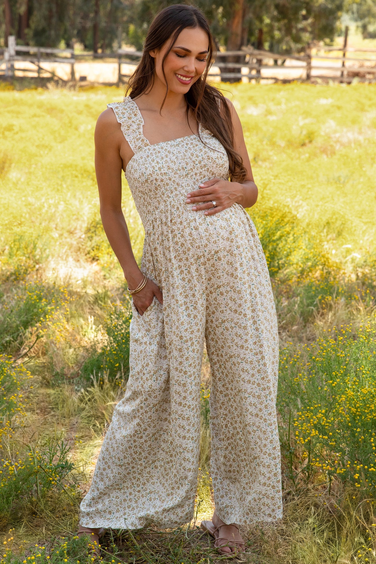 woman wearing a floral maternity jumpsuit with adjustable straps and pockets, posing with her baby bump in a field