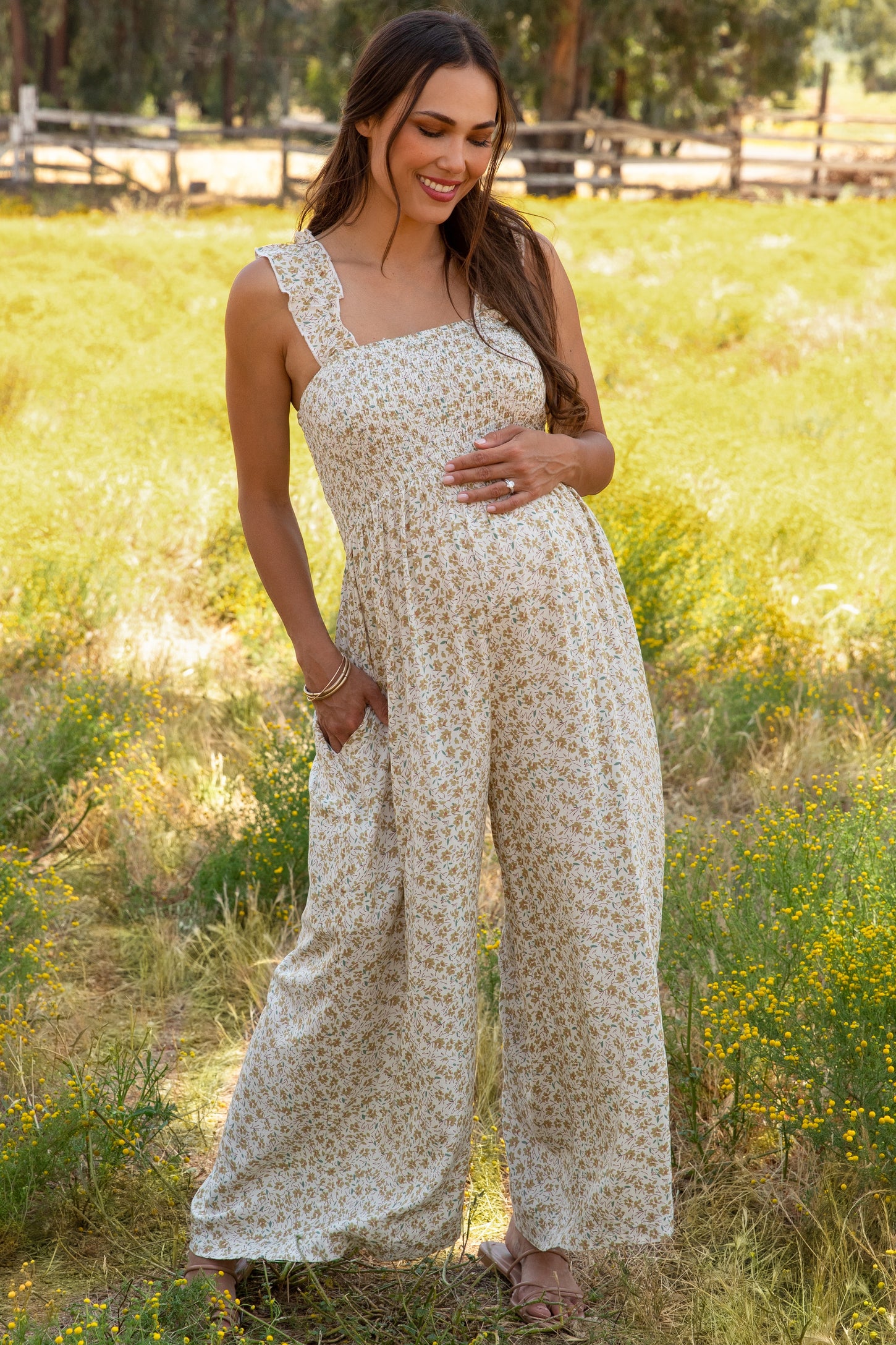 woman wearing a floral maternity jumpsuit with adjustable straps and pockets, posing with her baby bump in a field