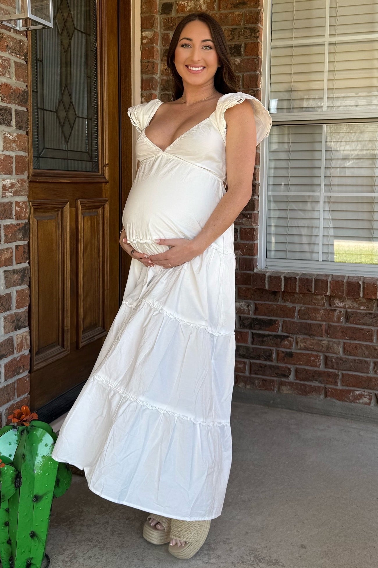 woman wearing a white tiered maternity dress and cradling her baby bump while standing at a front door