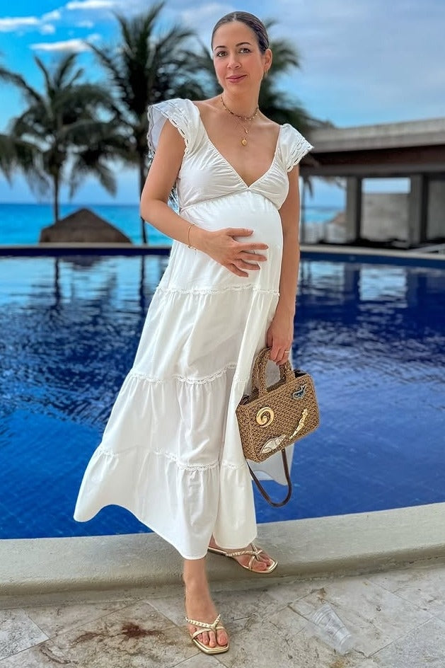 woman wearing a white tiered maternity dress, holding her baby bump, with a handbag, near a pool and palm trees in the background