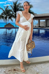 woman wearing a white tiered maternity dress, holding her baby bump, with a handbag, near a pool and palm trees in the background