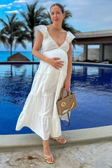 woman wearing a white tiered maternity dress and holding her baby bump by a pool with palm trees in the background