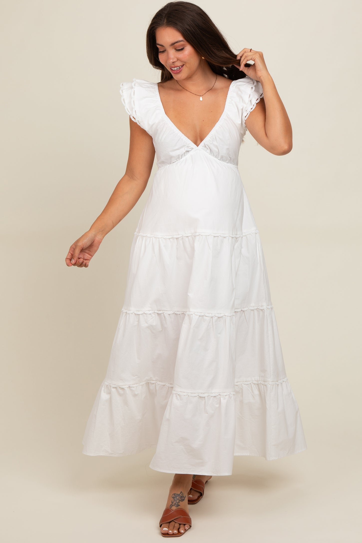 woman wearing a white tiered maxi maternity dress with ruffled sleeves and a plunging neckline, lightly swaying while walking
