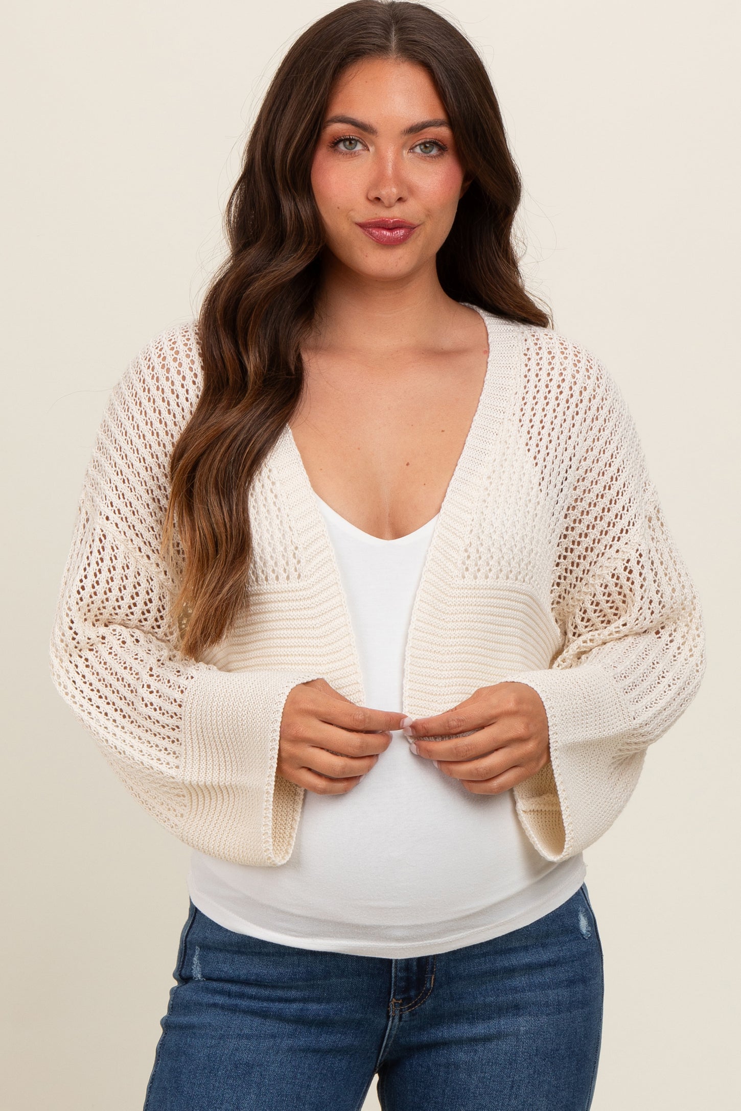 woman wearing a cream open-knit maternity cardigan over a white top and showing her baby bump