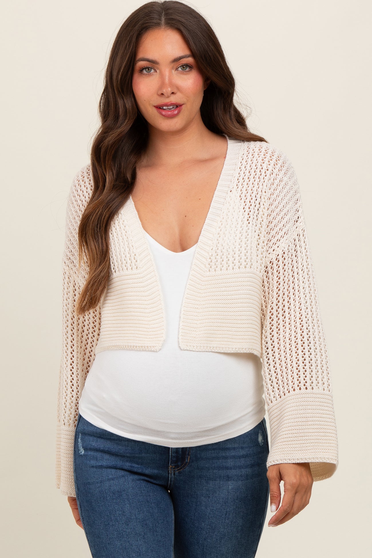 woman wearing a light beige knitted maternity cardigan over a white top with a visible baby bump and blue jeans