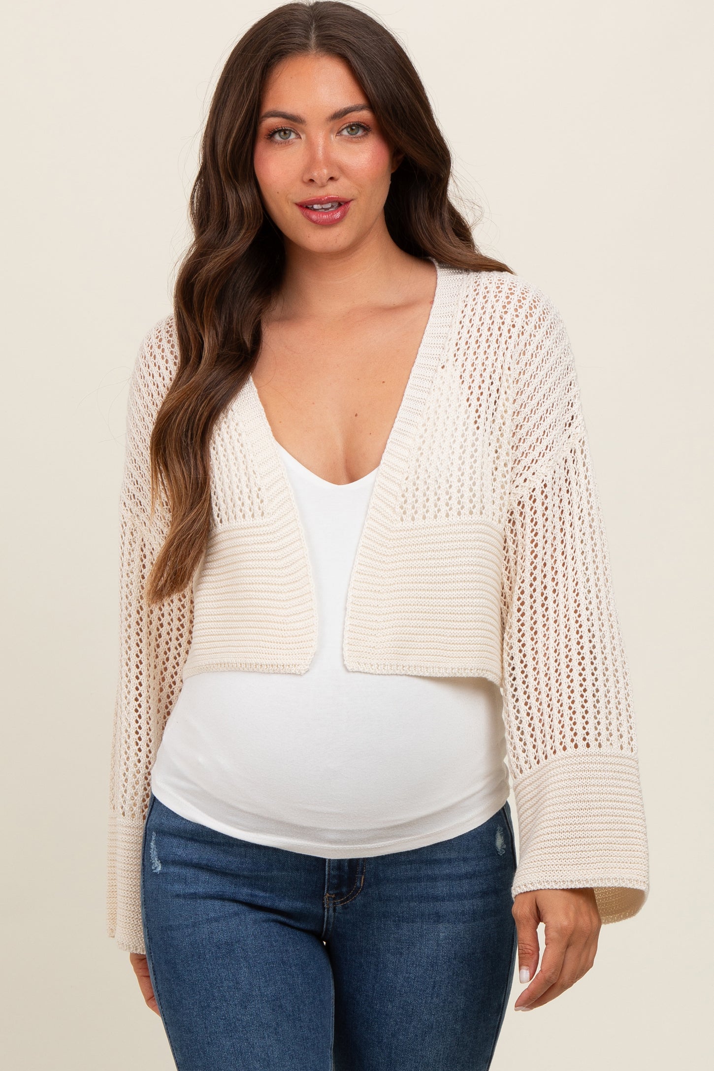 woman wearing a light beige knitted maternity cardigan over a white top with a visible baby bump and blue jeans