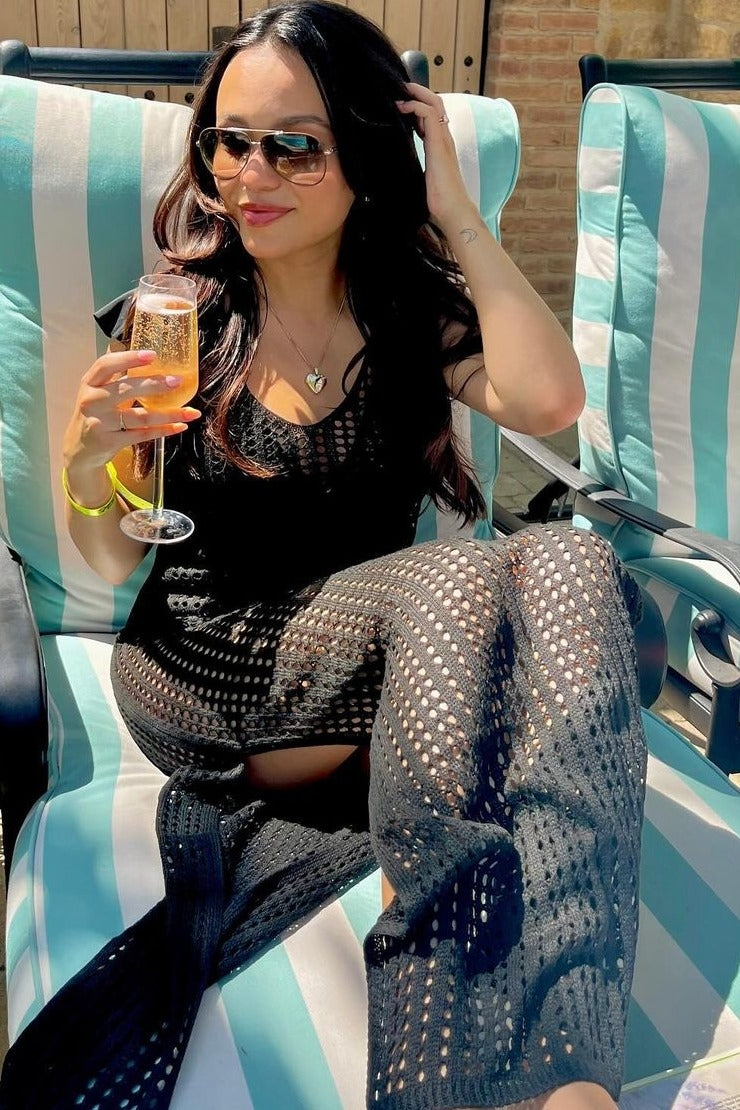 woman wearing a black crochet cover-up dress while sitting on a lounge chair and holding a drink