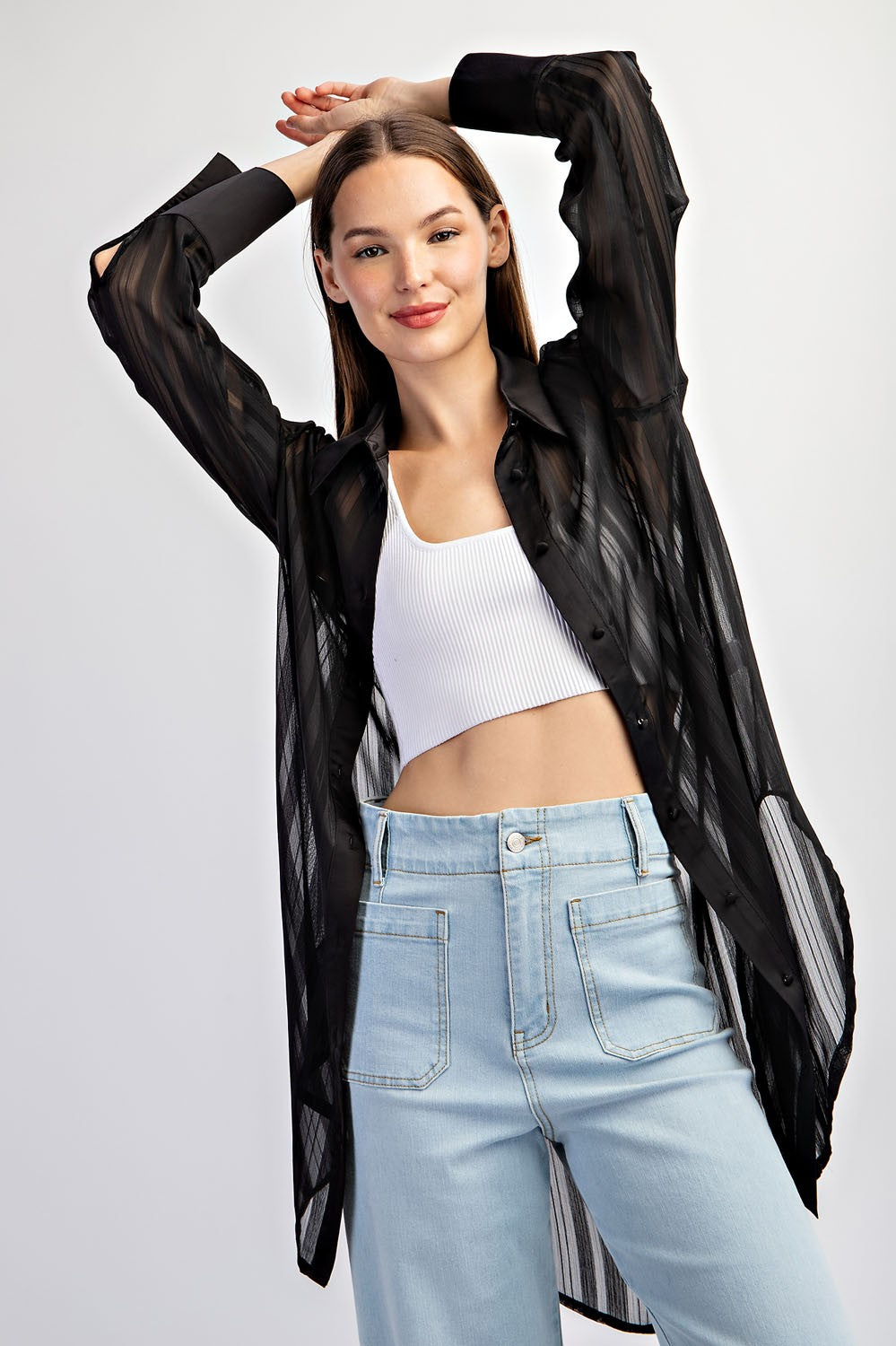 woman wearing a black sheer long sleeve button-up shirt over a white crop top and light blue high-waisted jeans